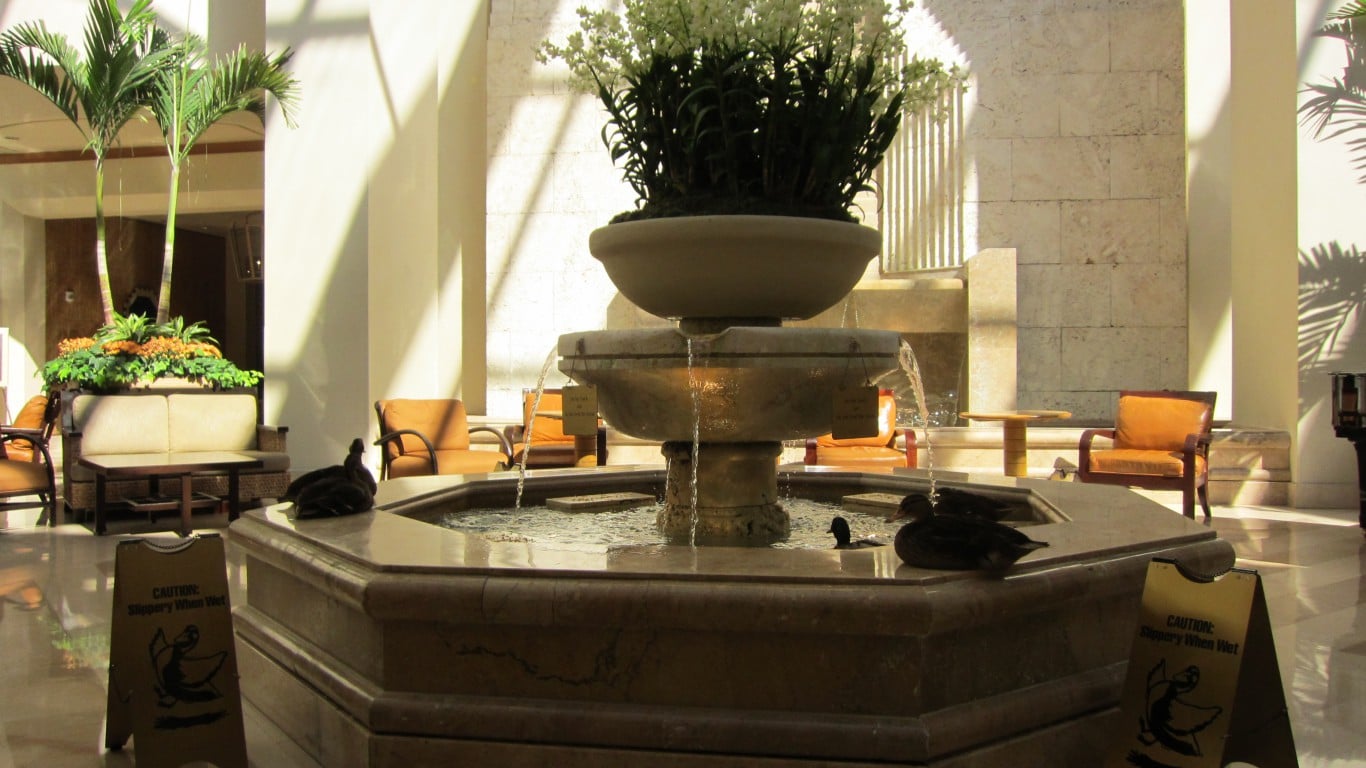A spacious, sunlit hotel lobby featuring a large, multi-tiered stone fountain with water flowing. White flowers and green plants adorn the top tier, while several dark-colored ducks are visible in the lower basin. Two yellow 'Caution: Slippery When Wet' signs are placed on the tiled floor near the fountain. In the background, there are comfortable beige sofas, orange armchairs, and tall palm trees in planters. The architecture includes white columns and stone-tiled walls, with strong shadows cast by sunlight.