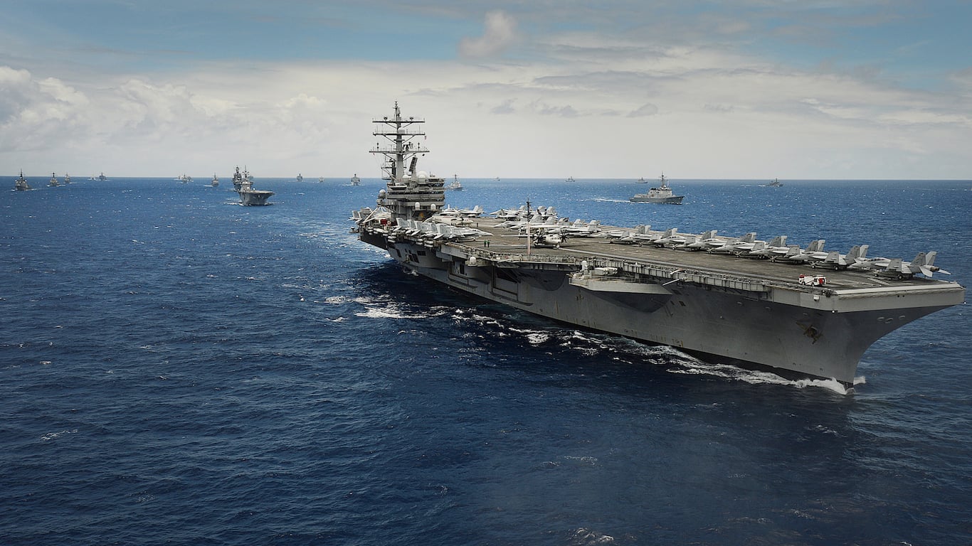 An aerial view captures a massive grey aircraft carrier, with dozens of fighter jets parked on its deck, cutting through deep blue ocean waters. Many smaller grey warships are visible in the distance, forming a large naval group under a partly cloudy blue sky, depicting a scene of vast military deployment.