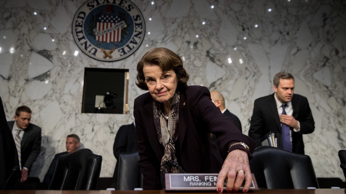 A candid, low-angle shot of Senator Dianne Feinstein, an older woman with brown hair, wearing a dark jacket and a patterned scarf, looking directly at the viewer. Her right hand, adorned with a ring and red nail polish, rests on a nameplate that reads 'MRS. FEIN RANKING MEMBER'. In the background, the circular United States Senate seal is visible on a light brown marbled wall, with several men in suits partially visible in the blurred background.