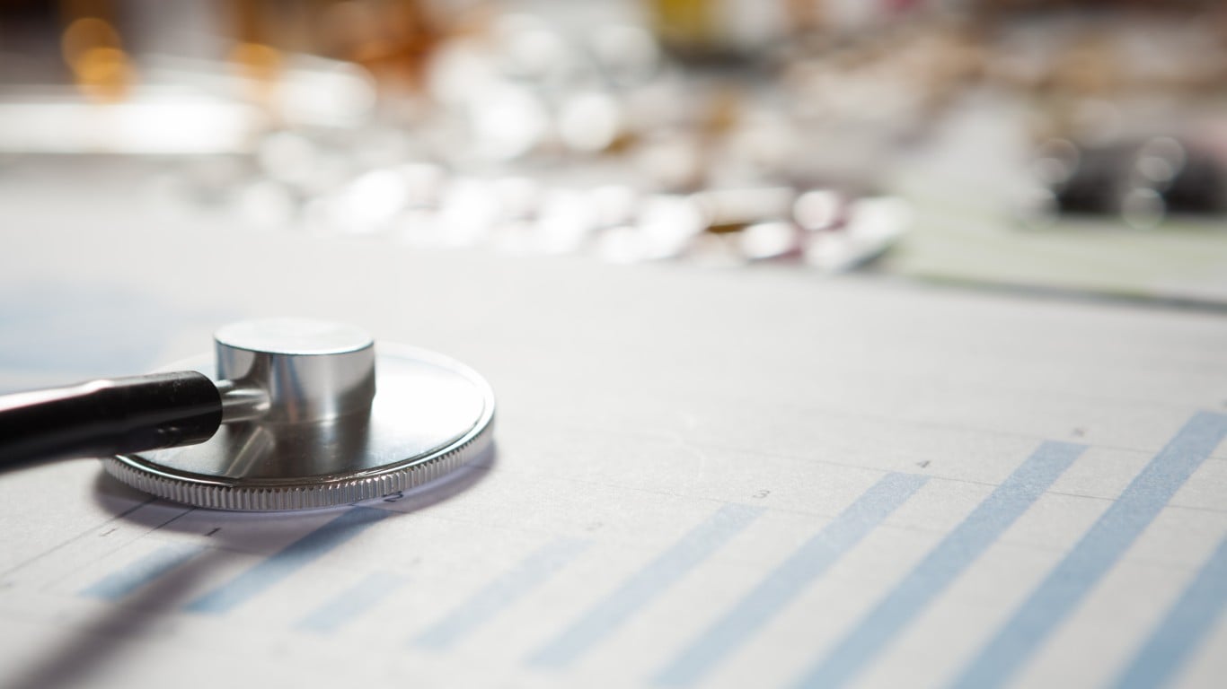 A close-up view of a silver and black stethoscope's chest piece resting on a white document that features light blue bar charts. The background is softly blurred with warm and bright bokeh lights, suggesting an office or medical setting.