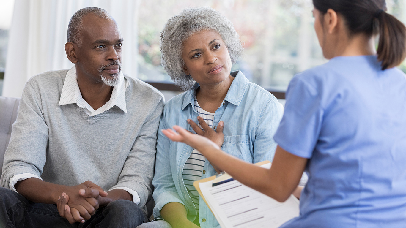 A Black couple sits opposite a healthcare professional, seen from the back, holding a clipboard. The woman, with gray curly hair, wears a denim shirt and has her hand on her chest, looking towards the professional. The man, with a gray beard and wearing a gray sweater, looks to the side. They are in a well-lit room, possibly a clinic or consultation office.