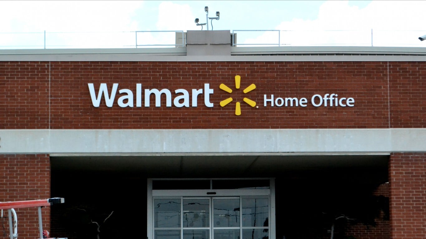 The exterior of the Walmart Home Office building, featuring a large white 'Walmart Home Office' sign with the yellow spark logo against a red brick wall. Below the sign are automatic glass entrance doors. The building has a light grey horizontal band above the entrance, and security cameras are mounted on the roof against a partly cloudy sky.