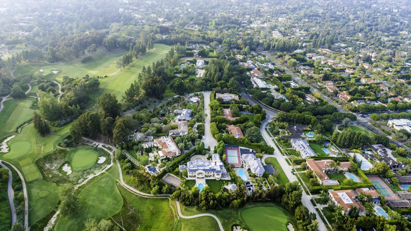 An aerial photograph shows a sprawling affluent residential neighborhood adjacent to a lush green golf course. Large, multi-story luxury homes with red tile roofs, swimming pools, and tennis courts are visible, surrounded by mature green trees and manicured lawns. A winding road connects the properties, and the golf course features greens, fairways, and sand traps under a soft, hazy sky.