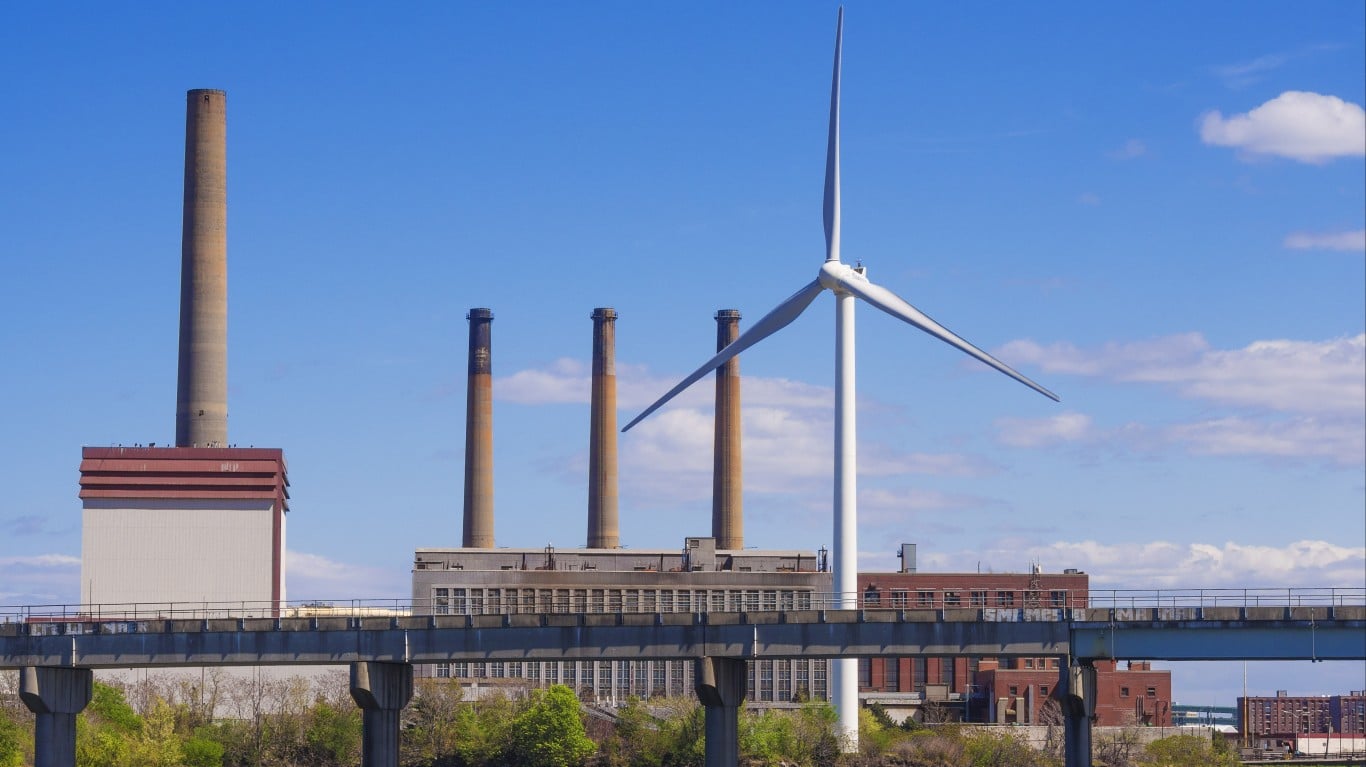 A wide shot shows a tall, light brown industrial smokestack on the far left, next to a white building with a red roof. To its right, an elevated concrete bridge with a dark railing spans across the midground. Behind the bridge, three more brown smokestacks rise from an industrial complex. A large, white wind turbine with three blades dominates the right side of the frame, its blades extended. The sky is clear blue with scattered white clouds.