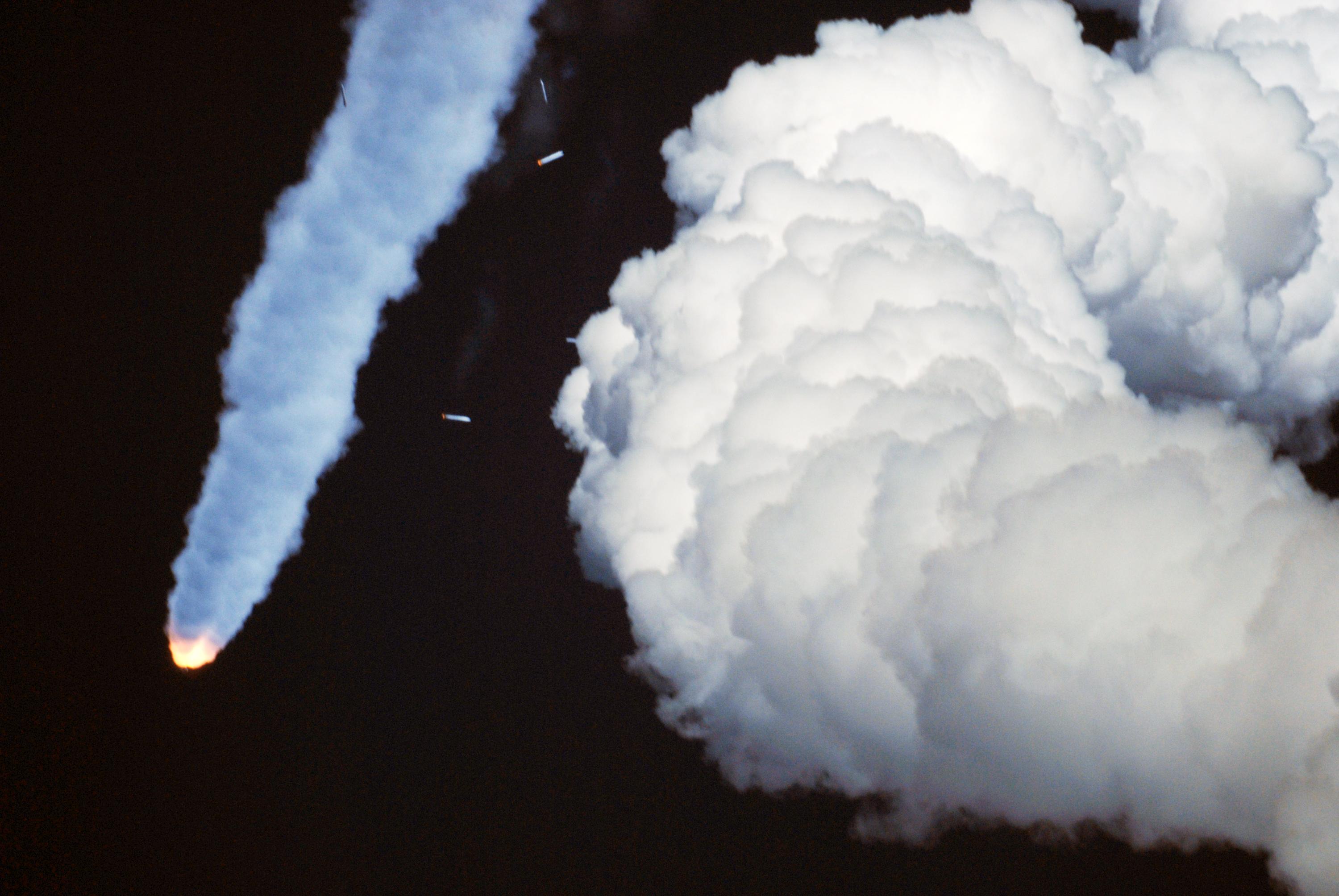 A powerful rocket launches against a dark background, with a bright orange flame at its base and a long, light blue exhaust plume extending upwards and to the left. Several smaller, glowing orange and white objects appear near the exhaust plume, possibly representing jettisoned stages or debris. To the right, a large, dense cloud of white and gray smoke fills the frame, likely from the launchpad.