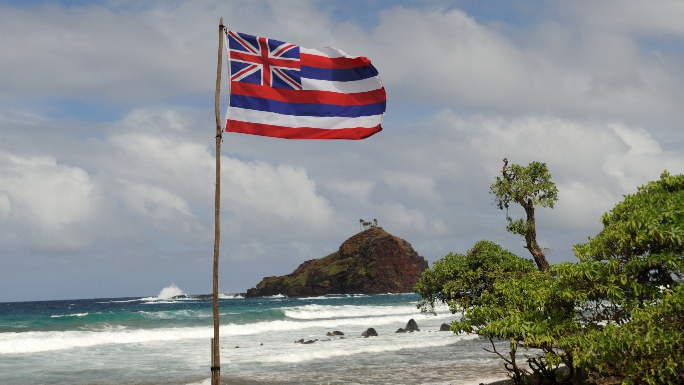 A Hawaiian flag flaps in the wind, with ocean waves and an offshore island in the background.