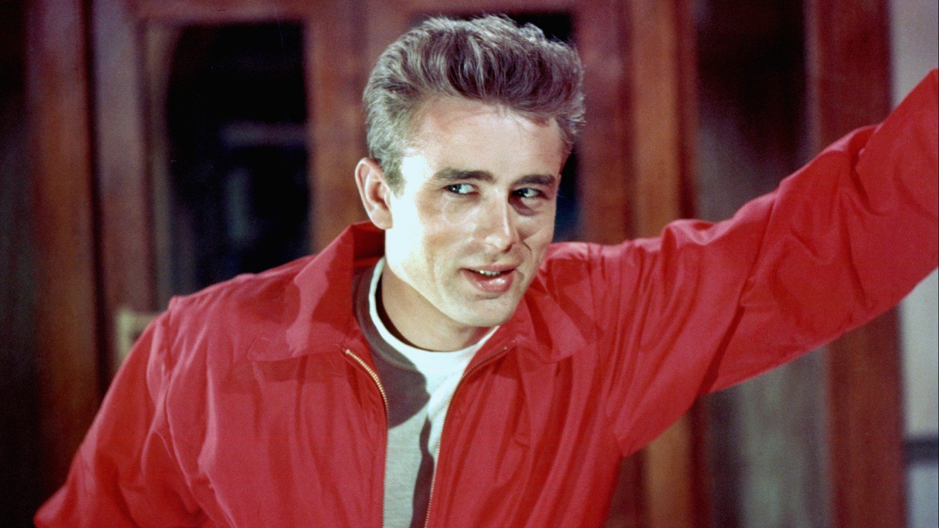 James Dean wears a white t-shirt and red jacket and leans against the wall. 
