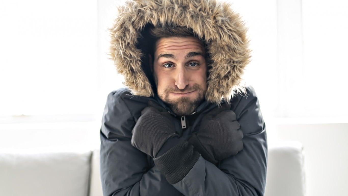 A man in a dark grey winter jacket with a brown fur-lined hood and black gloves shivers, pulling his arms into his chest. He has a beard and looks directly at the viewer with a slight frown and furrowed brow, conveying discomfort. The background is a bright, out-of-focus white.