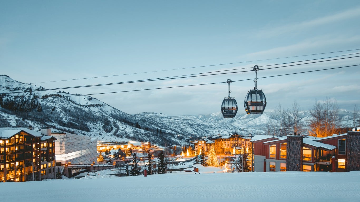A wide shot of a snow-covered ski resort town at dusk, under a pale blue, cloudy sky. Two dark gondola cars with internal lights are suspended on cables across the upper half of the frame. Below, multi-story buildings glow with warm yellow light from their windows, nestled among evergreen trees and bare deciduous trees. Snow-covered mountains rise in the background, and the foreground is a pristine white snowfield.
