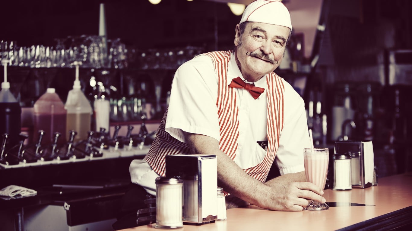 A smiling older man with a handlebar mustache, wearing a white cap and a red and white striped vest over a white shirt, leans on a diner counter. He holds a glass of pink milkshake in his left hand. The background is blurred, showing a dark diner interior with various chrome soda fountain equipment and syrup bottles.