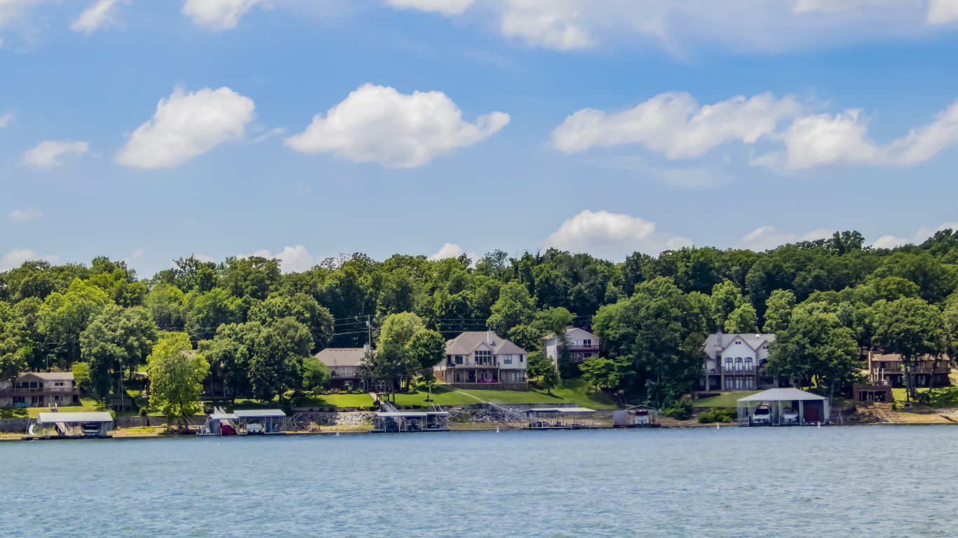 A wide shot of several large, well-maintained homes situated on a green, tree-covered hillside overlooking a serene blue lake. Each house has a private boat dock or boathouse extending into the calm water. The sky above is bright blue with scattered white cumulus clouds.