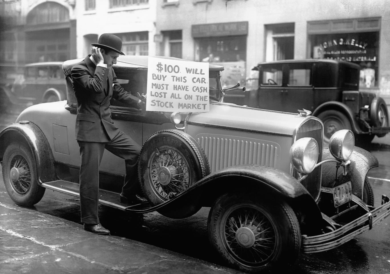 A black-and-white photograph shows a man in a suit and bowler hat standing beside an antique car. He is looking down, with one hand near his ear and the other resting on the car's open window. A handmade sign is attached to the windshield, stating: '$100. WILL BUY THIS CAR. MUST HAVE CASH. LOST ALL ON THE STOCK MARKET.' The car is a polished, older model with wire-spoke wheels, parked on a wet street. Other cars and buildings are blurred in the background, suggesting an urban setting.