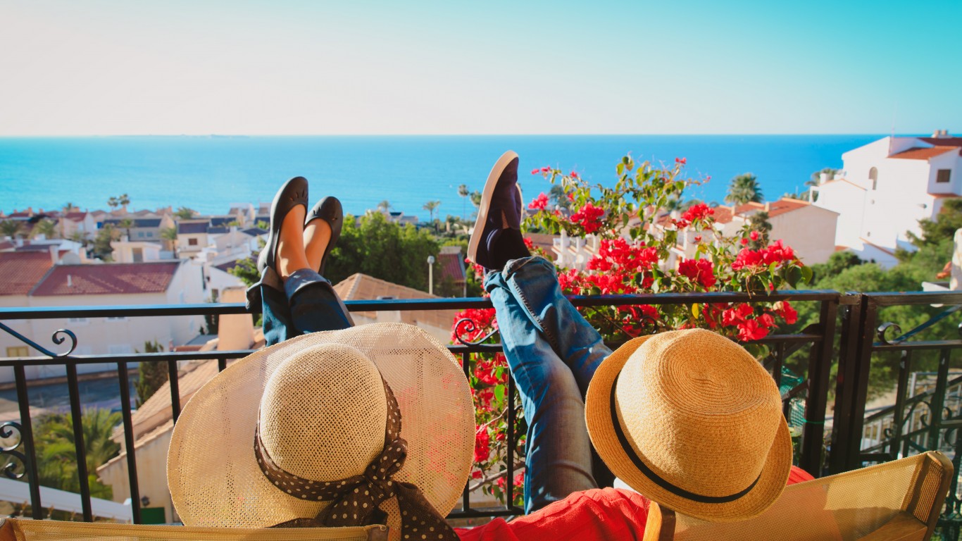 A high-angle view from behind shows two people relaxing in chairs on a balcony, with their feet propped up on the black metal railing. The person on the left wears a wide-brimmed straw hat with a patterned band and dark flat shoes. The person on the right wears a smaller straw hat, a red shirt, blue jeans, and dark shoes. Beyond the balcony, a coastal town with white and terracotta-roofed buildings spreads across a hillside, leading to a vast, calm blue ocean under a clear sky. Bright red bougainvillea flowers are visible near the railing.