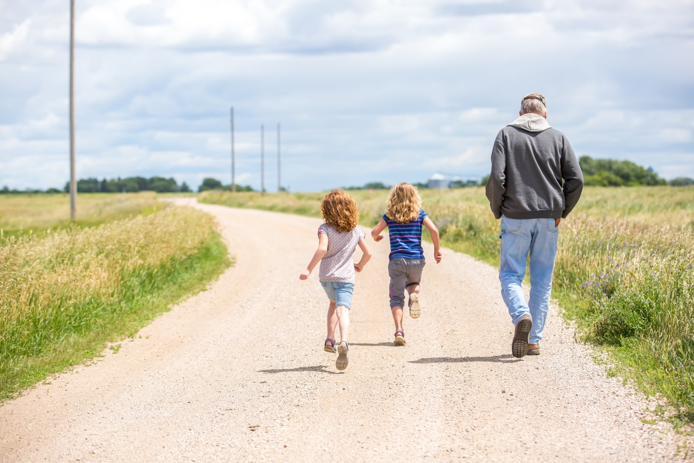 Two young girls run on a gravel road beside a walking older man. 