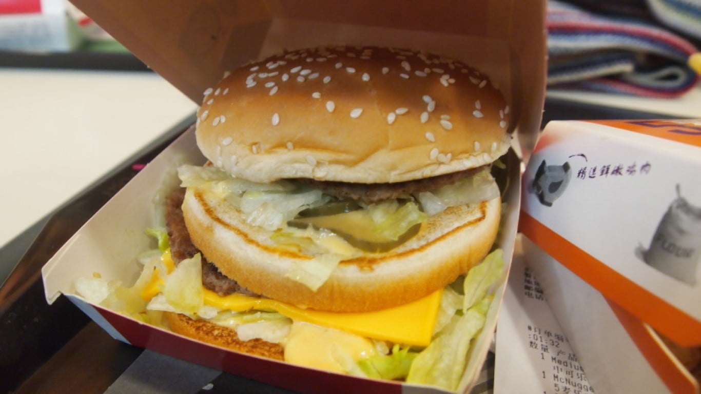 A close-up, slightly elevated view of a McDonald's Big Mac inside its paper box. The burger features a golden-brown sesame seed bun, two meat patties, melted yellow cheese, shredded green lettuce, and sliced pickles. To the right, part of a white and orange McDonald's receipt and another branded package are visible, slightly out of focus.