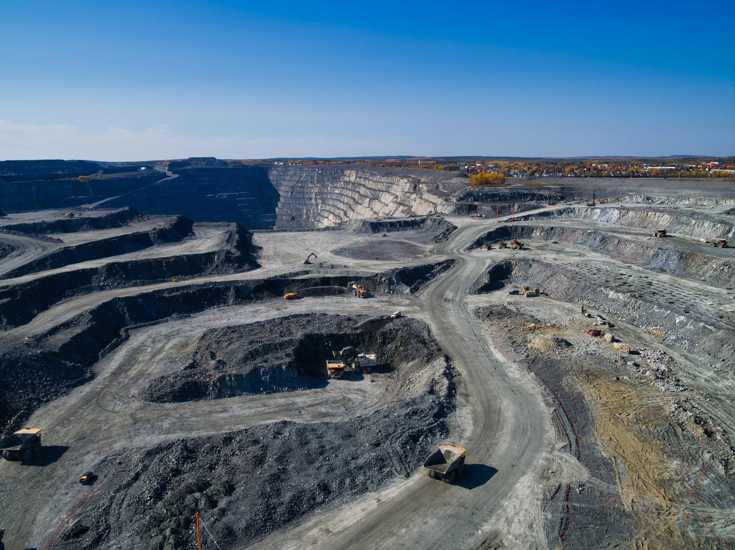 An aerial wide shot of a vast, multi-level open-pit mine under a clear blue sky. Dark grey and light grey rock formations define the terraced walls and winding dirt roads where several yellow heavy excavators and large dump trucks are visible. In the distance, a small town with colorful autumn trees can be seen on the horizon above the quarry.