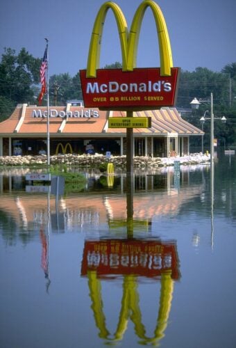 A McDonald's restaurant and its prominent golden arches sign stand partially submerged in calm floodwaters under a clear blue sky. Sandbags are visible around the base of the brick building, and a smaller sign on the main pole reads 'OPEN WATERFRONT DINING.' The entire scene, including an American flag on a pole, is reflected clearly in the water.