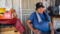 A medium shot of an older man with dark skin, wearing a blue short-sleeved t-shirt and a black bucket hat with a light-colored logo. A grey and black towel is draped around his neck. He is seated, looking downwards and slightly to his right. Behind him is a white metal security door and a textured, light-colored wall with peeling paint. To his right, a black oscillating fan is visible, and to his left, cardboard boxes for Sunkist Cara Cara oranges are stacked.