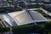 An aerial shot of a large, contemporary building in an urban setting. The building has a distinctive multi-faceted roof covered in both light-colored metal panels and numerous dark solar panels. A prominent angular glass facade on one side also features integrated solar panels. The building is surrounded by dense green trees and other urban buildings are visible in the background under a clear sky.