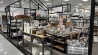 A wide shot of a brightly lit retail store aisle showcasing a home goods display. A prominent black, house-shaped frame structure with 'Hearth & Hand with Magnolia' signage stands over shelves filled with white and grey dishware, wooden bowls, and boxed items. Further into the aisle, tables display various kitchenware, mugs, and decorative elements, while woven baskets contain neatly folded blankets and towels. The overall setting is clean, organized, and modern with a neutral color palette.