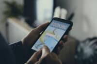 A close-up shot shows a person holding a black smartphone horizontally with both hands, focusing on the screen. The phone displays a travel booking application with the word 'BOOKING' prominently at the top. Below it is a white search bar with a magnifying glass icon. The background image on the app screen features a yellow inflatable ring, a blue floating object, and a person's legs wearing green flip-flops, suggesting a vacation or travel destination.