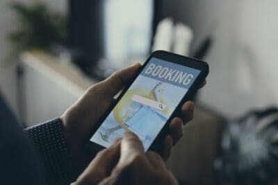 A close-up shot shows a person holding a black smartphone horizontally with both hands, focusing on the screen. The phone displays a travel booking application with the word 'BOOKING' prominently at the top. Below it is a white search bar with a magnifying glass icon. The background image on the app screen features a yellow inflatable ring, a blue floating object, and a person's legs wearing green flip-flops, suggesting a vacation or travel destination.