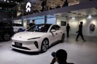 A light pink Nio ET5 electric car is parked on a grey tiled floor, prominently displayed at an indoor auto exhibition. Several people are visible in the background and foreground, including one person leaning towards the car's windshield and another person walking past a white wall featuring "Clean" and "NIO Power" signs. A photographer with a large camera lens is seen from behind in the lower right corner. Above, a second floor with seating and a large white Nio logo is visible.