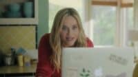 A blonde woman in a red long-sleeved shirt looks wide-eyed and shocked at a silver laptop. Her mouth is slightly open, conveying surprise. The background is a home setting with yellow patterned kitchen tiles on the left and a blurred window on the right.