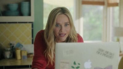 A blonde woman in a red long-sleeved shirt looks wide-eyed and shocked at a silver laptop. Her mouth is slightly open, conveying surprise. The background is a home setting with yellow patterned kitchen tiles on the left and a blurred window on the right.