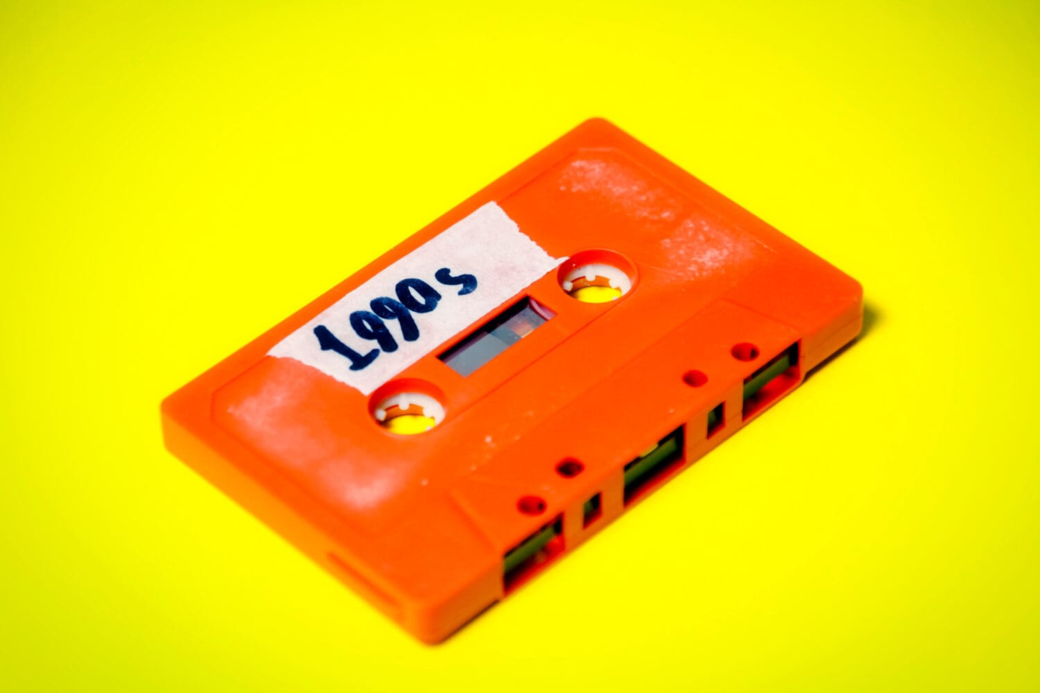 A vintage cassette tape (obsolete music technology), orange on a yellow surface, angled shot, carrying a label with the handwritten text 1990s.