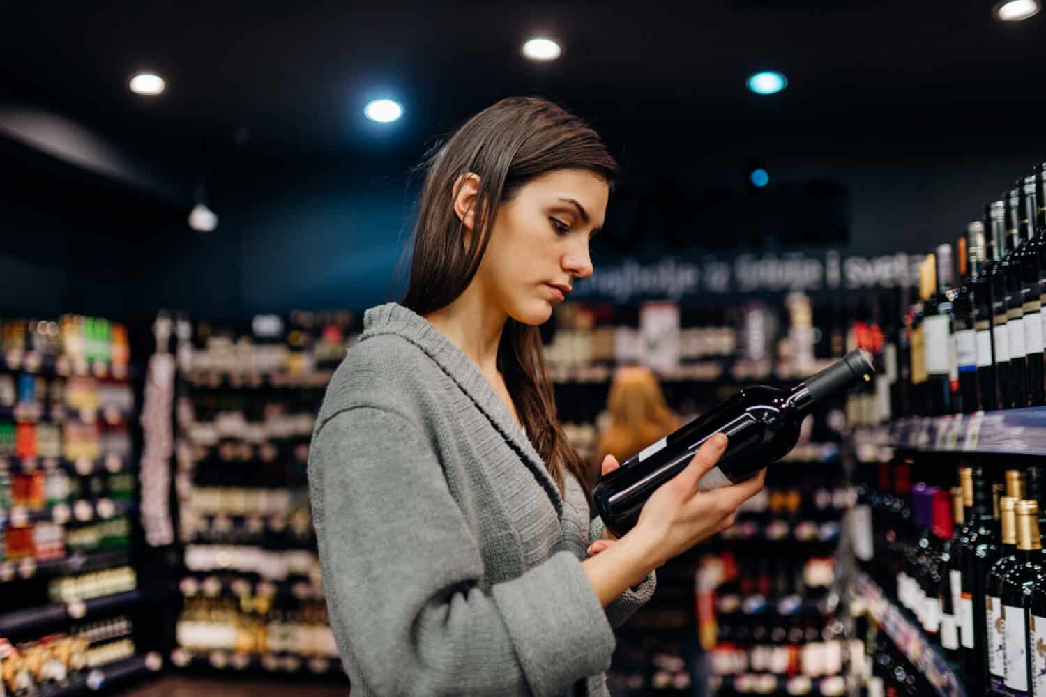 Woman shopping for expensive wine in supermarket alcohol store.Choosing and buying good cheap wine.Benefits of drinking wine.Resveratrol.Everyday binge drinking.Oenology.Mediterranean diet.Alcoholism