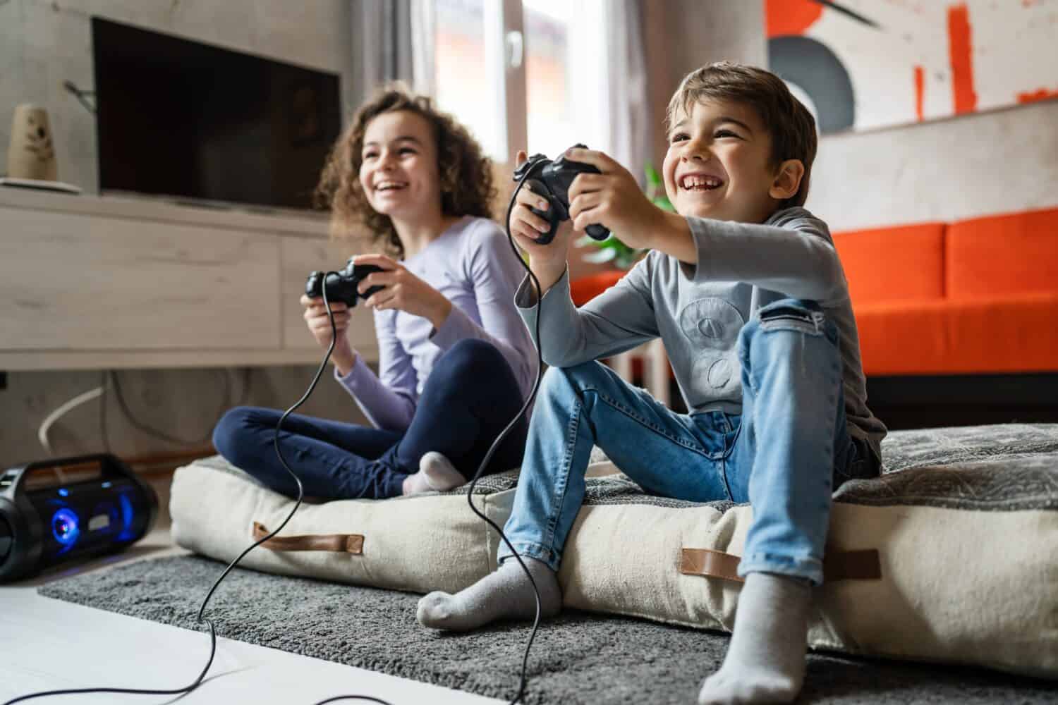 Two children small caucasian brother and sister happy children siblings boy and girl playing video game console using joystick or controller while sitting at home real people family leisure concept