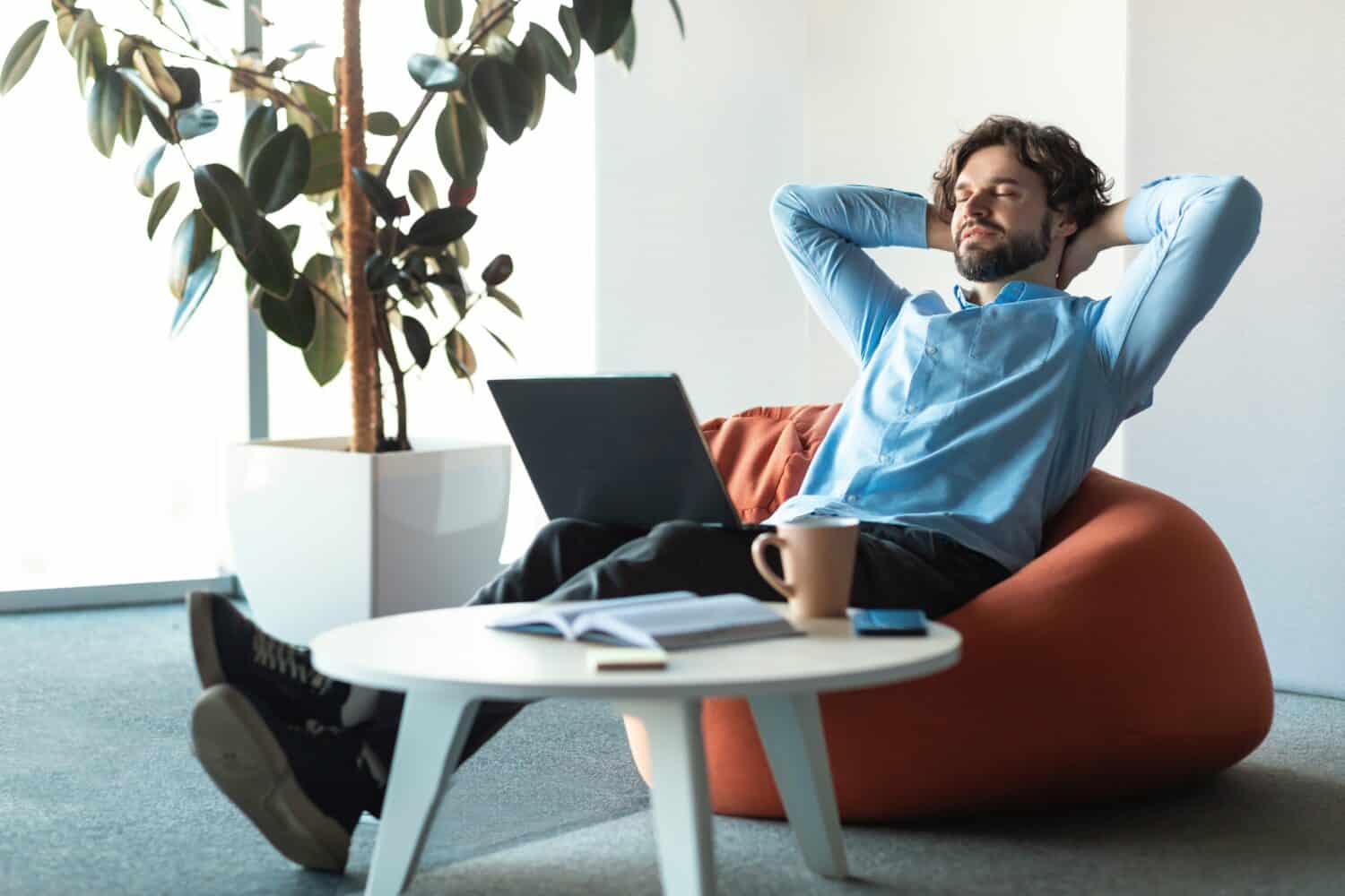 Taking Break From Work. Smiling business man relaxing on beanbag sitting on pouf chair and resting, happy male leaning back at workplace, cheerful guy with closed eyes holding hands behind head