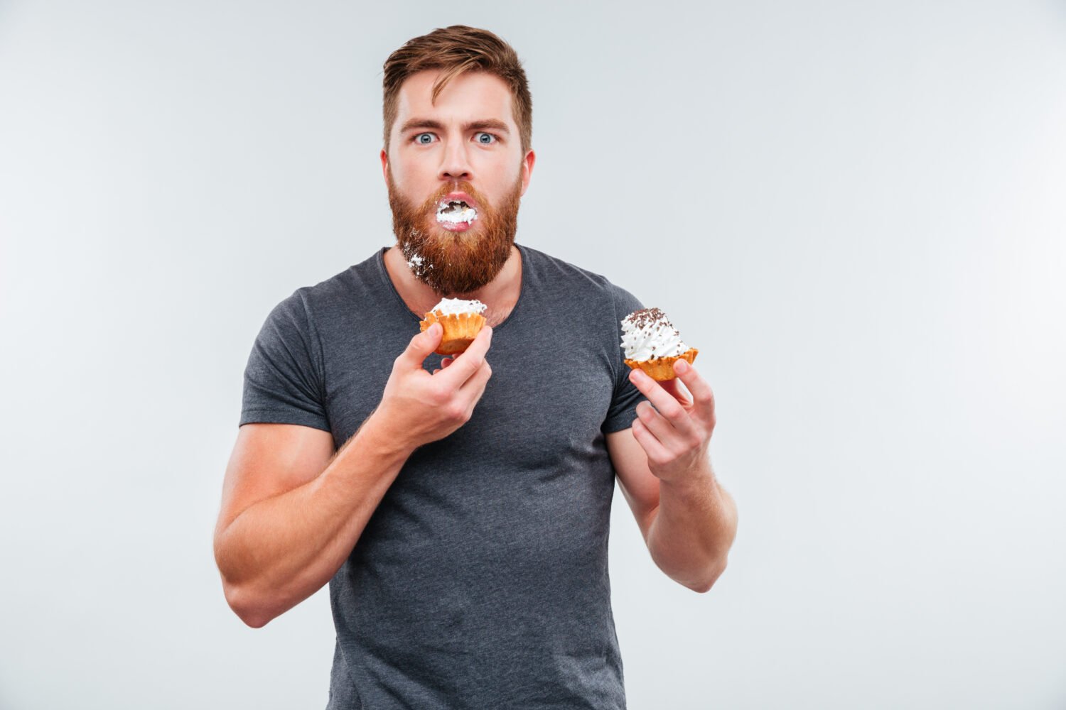Filthy bearded young man eating cream cakes isolated on white background
