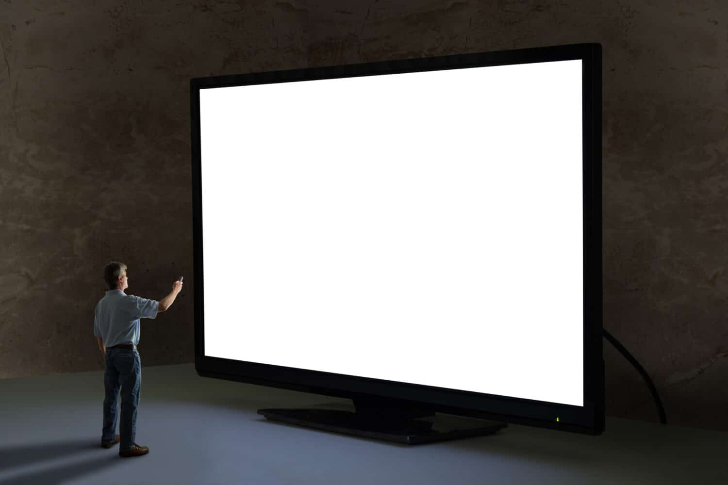 Man pointing tv remote control at world's biggest television towering over him shining bright light in a dimly lit room with clean blank screen for your message and image sending a powerful message