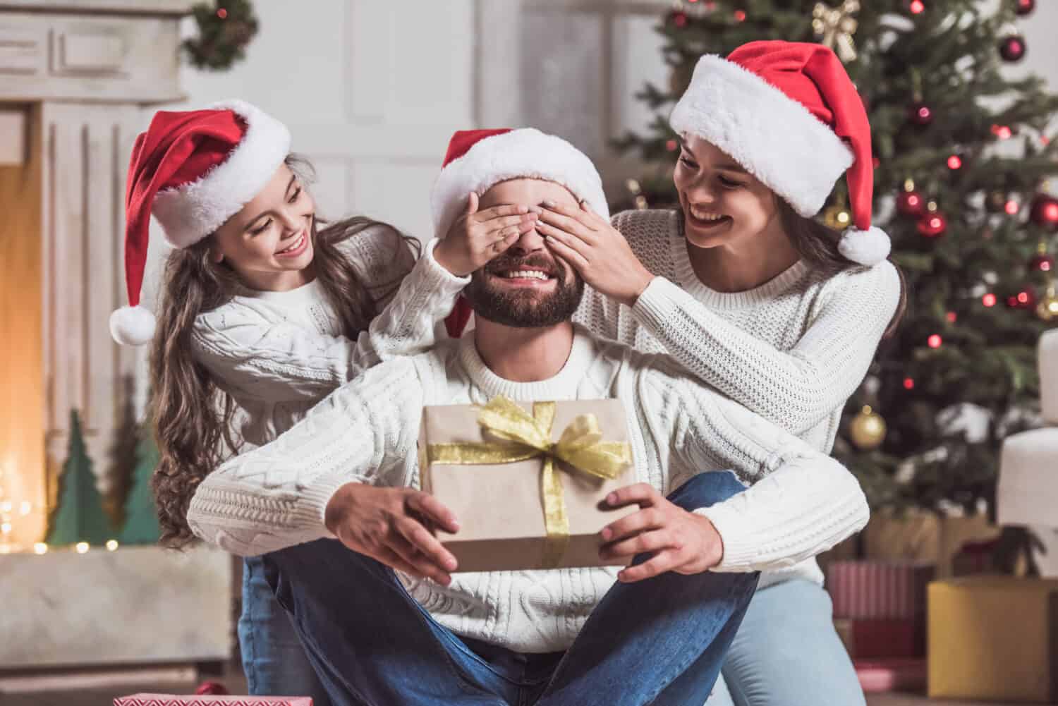 Merry Christmas and Happy New Year! Handsome man is holding a present and smiling while his daughter and wife covering his eyes