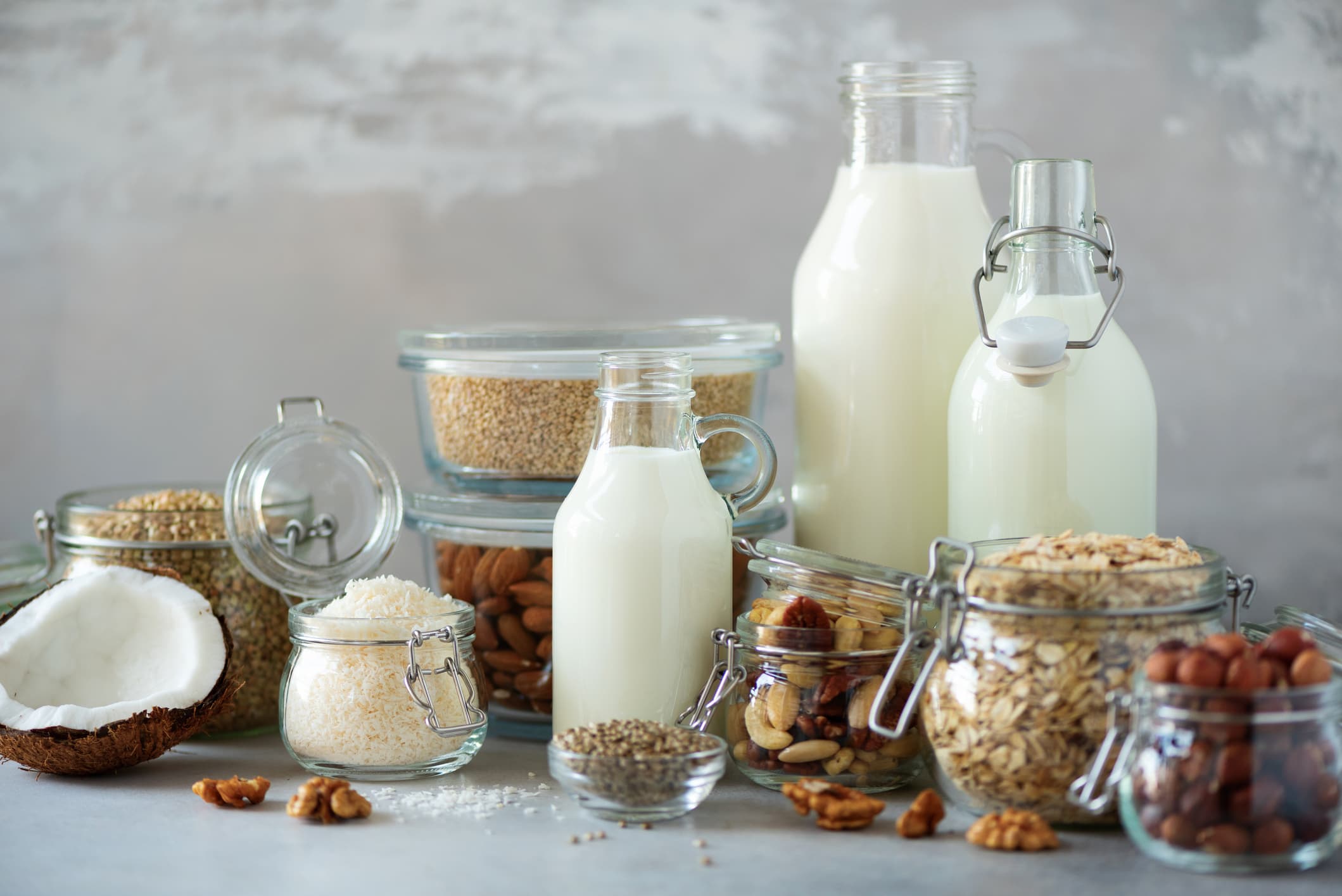 Glass bottles of vegan plant milk and almonds, nuts, coconut, hemp seed milk on grey concrete background. Banner with copy space. Dairy free milk substitute drinks and ingredients