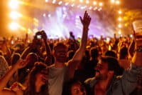 A dynamic, low-angle shot of a diverse crowd enjoying a live concert at night. Bright orange and purple stage lights illuminate the smiling faces and raised hands of audience members, some holding drinks or cameras. The mood is joyful and energetic, with visible motion and interaction among the attendees.
