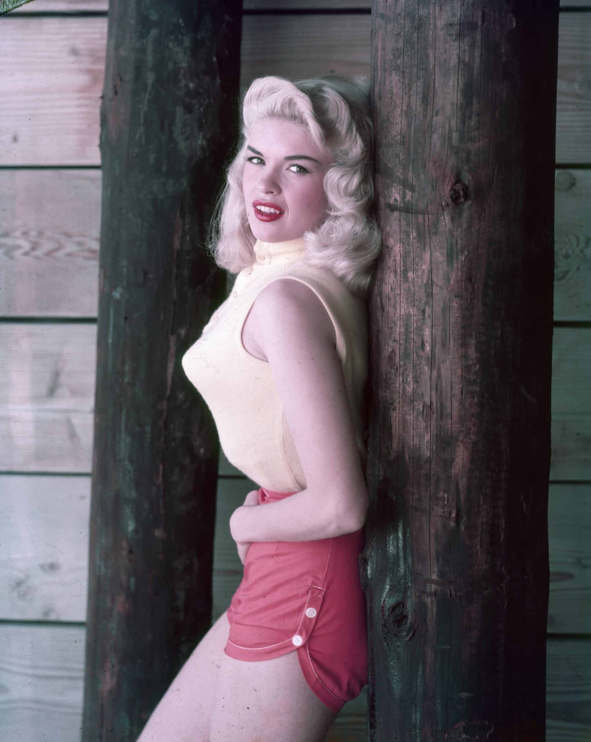 American actor Jayne Mansfield wears a sleeveless sweater, pointy bra and red shorts while leaning against a wooden post. 