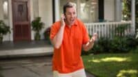 A man with short brown hair, wearing a bright orange polo shirt and light-colored pants, stands outdoors in front of a house. He holds a black cell phone to his left ear, his mouth wide open in an expressive shout or laugh, and his right hand is raised and open in a gesturing motion. In the background are a brown front door, potted plants, a white porch railing, and green grass.
