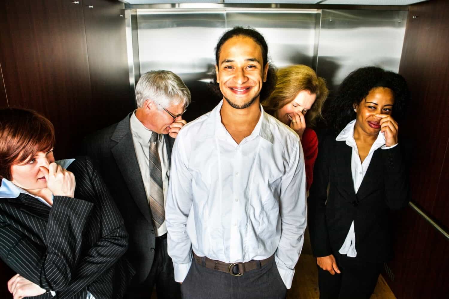 A young man smiles in embarrassment in an elevator while people hold their noses.