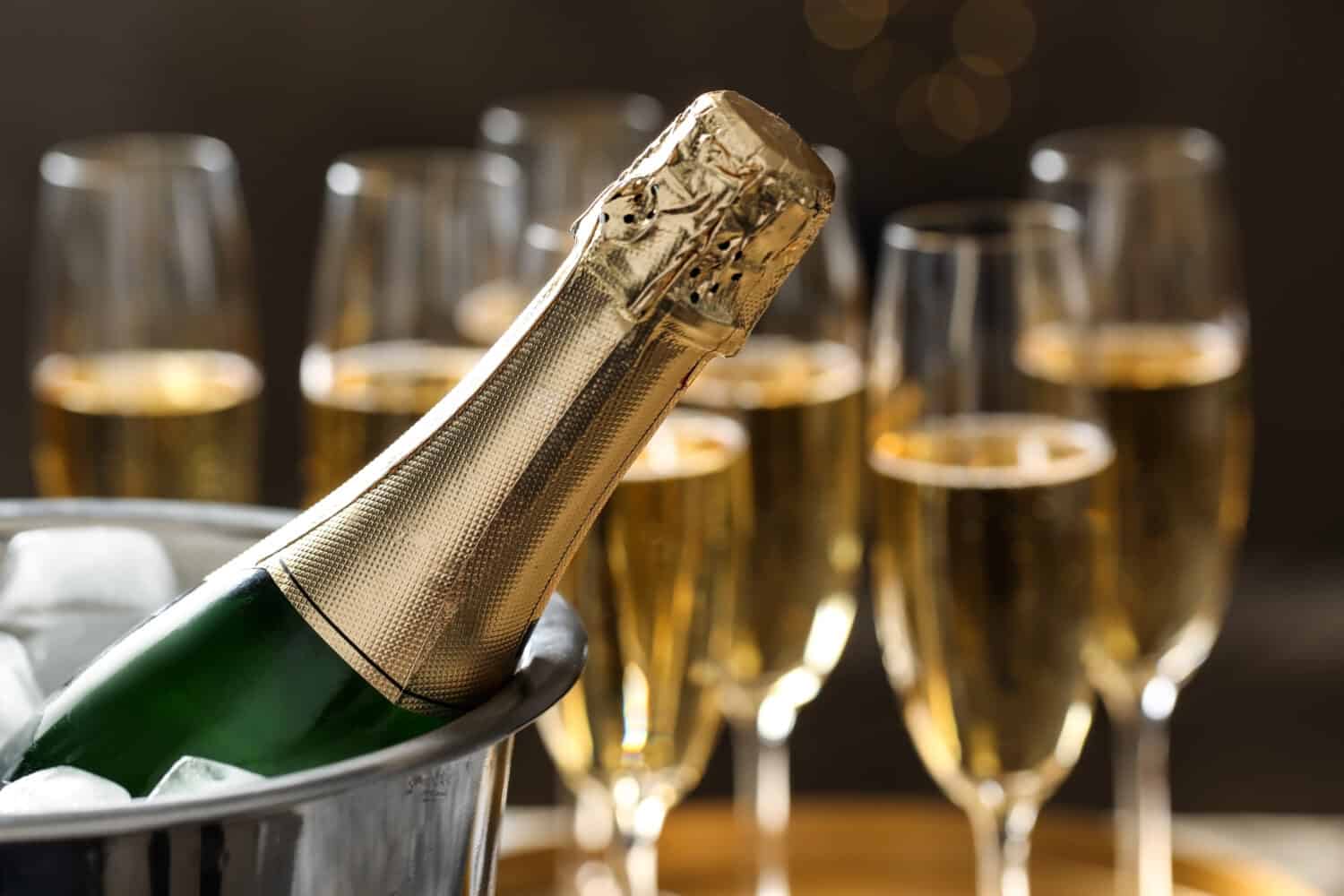 Bottle of champagne in bucket with ice and glasses on blurred background, closeup. Space for text