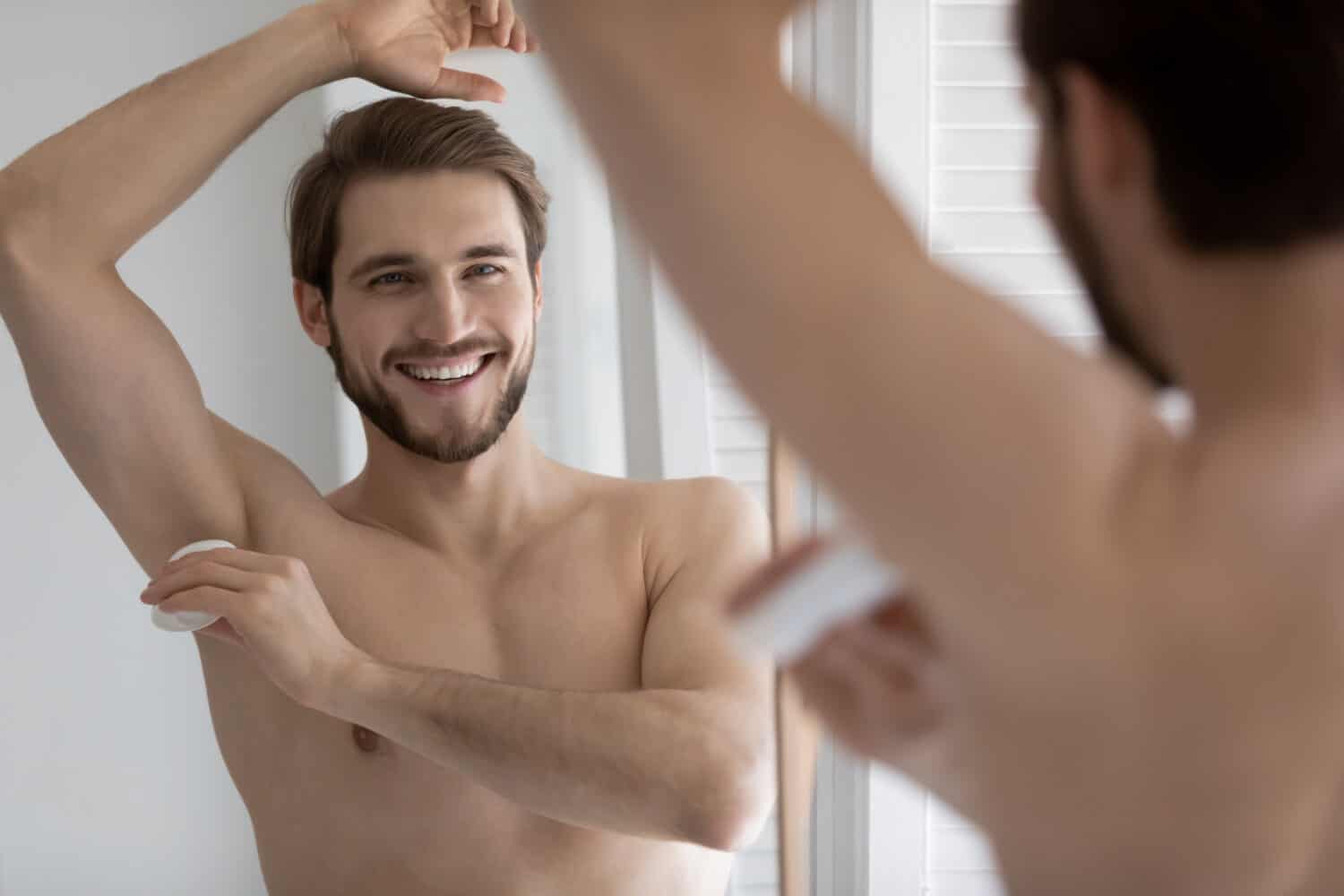 Mirror reflection smiling young man applying antiperspirant on armpit after shower, standing in bathroom, satisfied handsome guy enjoying morning routine procedure, using stick underarm deodorant