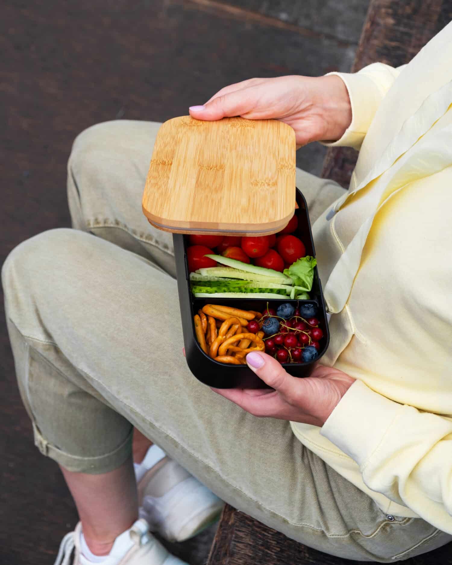 Young woman having a snack with healthy food in lunch box during the walk. Lunchbox, eco cutlery, reuseable dish, bamboo cutlery. Healthy lunch outdoors
