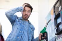 A man with dark hair and a beard, wearing a denim jacket over a light blue t-shirt, stands at a gas pump. He holds a green and black fuel nozzle in his right hand and looks directly at the viewer with a wide-eyed, open-mouthed expression of shock, with his left hand on his head. A gas pump display is visible in the bright background.