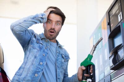 A man with dark hair and a beard, wearing a denim jacket over a light blue t-shirt, stands at a gas pump. He holds a green and black fuel nozzle in his right hand and looks directly at the viewer with a wide-eyed, open-mouthed expression of shock, with his left hand on his head. A gas pump display is visible in the bright background.