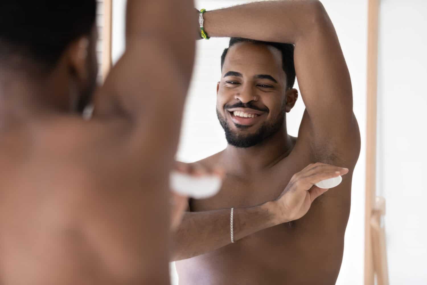 Calm handsome young well built man using deodorant, applying antiperspirant on underarm armpit skin after shower in bathroom at mirror, caring for hygiene, preventing sweating. Bath routine concept