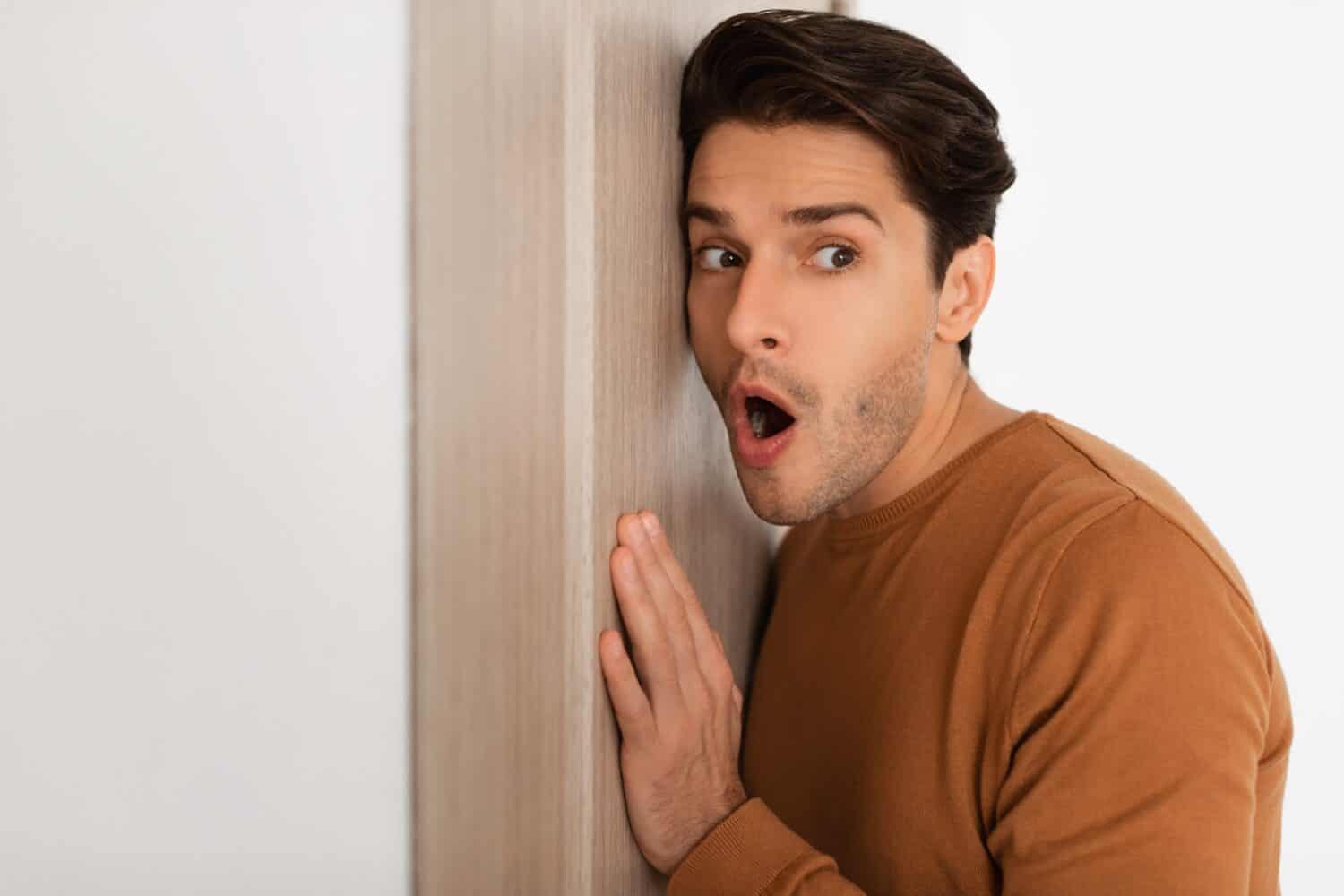 Closeup portrait of amazed man eavesdropping on private conversations, spying and listening through the door with a shocked expression at what he has overheard, man snooping leaning on wall