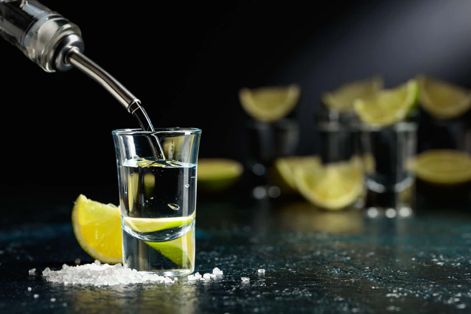 Tequila is poured into a glass. Tequila shots with lime slices and sea salt on a dark blue table.