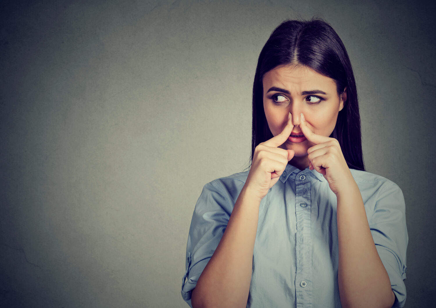Woman pinches nose with fingers looks with disgust away something stinks bad smell isolated on gray background.